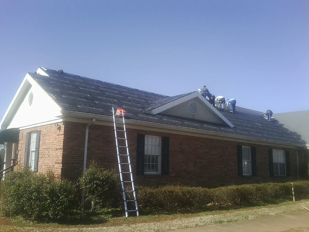 Crew tearing shingles off a brick ranch roof with a ladder set — Metal Roof Repair work by our crew in Tracys Landing