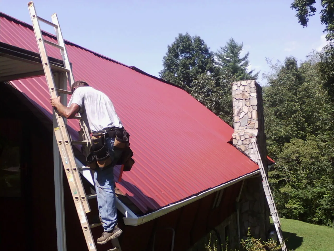 Roofer on a ladder beside a red metal roof and stone chimney — Soffit & Fascia Repair by Roofing Brothers in Tracys Landing
